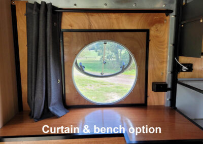 Caravan interior showing a comfortable upholstered bench seating area with a small wooden table and curtain, ideal for dining or lounging.