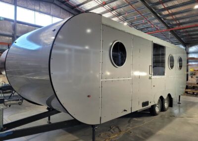 Front angled view of a silver Probond caravan with two round windows and a large rectangular window, inside Ballarat factory.