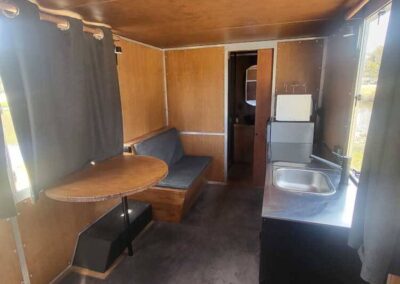 Caravan interior showing a cozy dining nook with a small round table and bench seating, leading to a compact kitchen area.