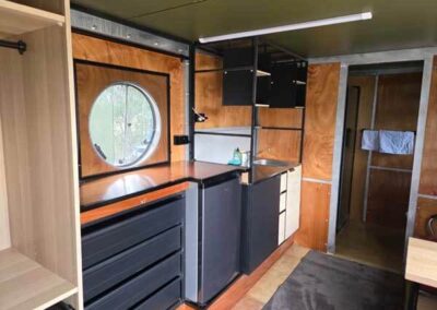 Caravan kitchen and living space featuring a small refrigerator, sink, open shelving, and a porthole window with wooden accents.