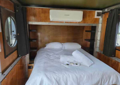 Caravan bedroom with a queen-sized bed, white bedding, and overhead air conditioning unit, featuring wooden walls and black accents.