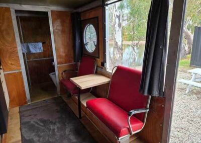 Caravan interior with two red retro-style seats and a small table, looking towards a bathroom door and a window with outdoor views.