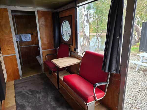 Caravan interior with two red retro-style seats and a small table, looking towards a bathroom door and a window with outdoor views.