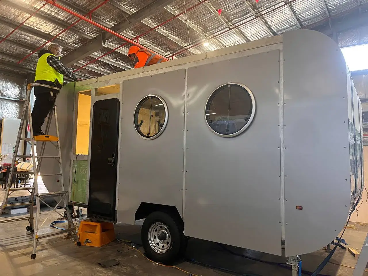 Workers constructing a 20ft caravan with a light grey exterior and round windows, one on a ladder, in a Ballarat Hammervan facility.