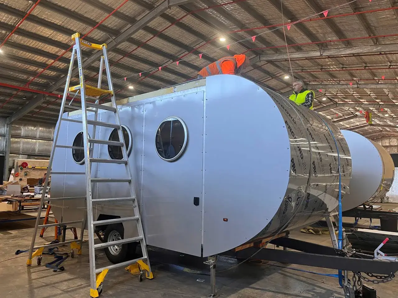 Two workers on ladders assembling the roof and side panels of a 20ft modern caravan with round windows in Ballarat factory.