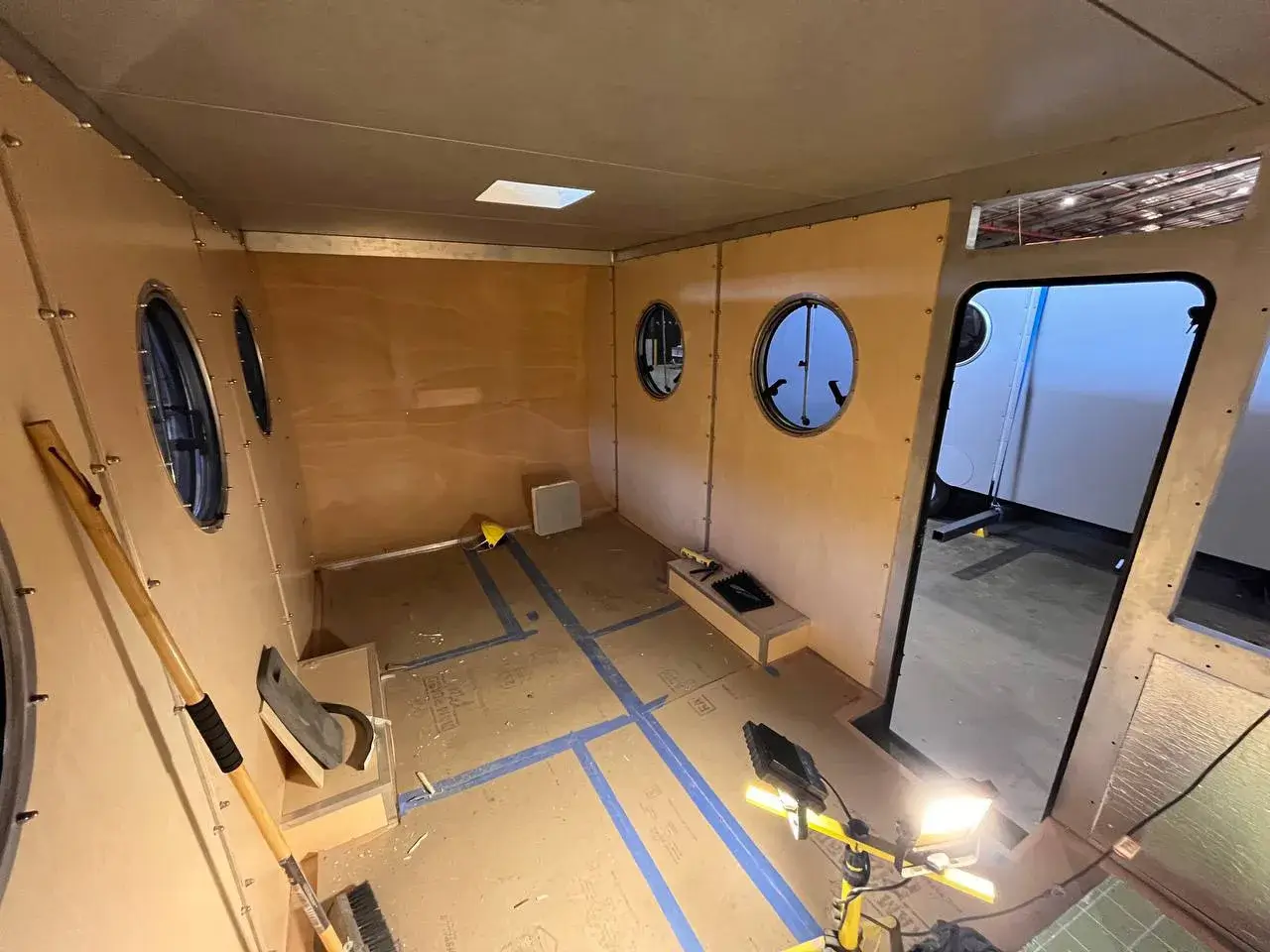 Interior view of a 20ft caravan under construction, showing plywood walls, round windows, and the floor with blue markings.